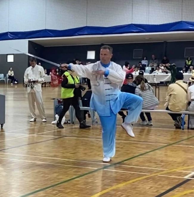 Peter Davey balanced on one leg with the straight sword extended behind him, competing in blue and white silk
